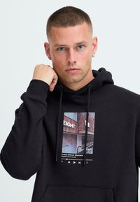 Joven con cabello corto y pendiente lleva una sudadera negra con una fotografía urbana en el frente y íconos de reproductores de música.