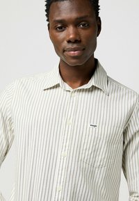 Camicia a righe bianca e beige con colletto, dotata di taschino sul petto e di un piccolo logo. Tessuto morbido con vestibilità su misura.