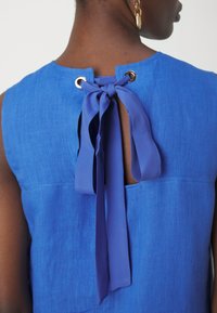 Haut en lin bleu avec un col rond, doté d'un dos ajouré fermé par un lien en tissu et des œillets dorés. Texture lisse avec une coupe décontractée.