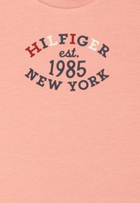 Ljusrosa bomullströja med marinblå och röda tryckta texter som säger "HILFIGER est. 1985 NEW YORK" i en cirkulär design. Enkel och slät textur.