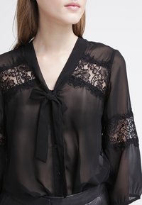 Czarna przezroczysta bluzka z koronkowymi akcentami na rękawach i wiązaniem przy dekolcie, zapinana na guziki z gładką fakturą.