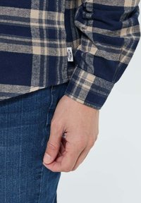 Chemise en flanelle à carreaux bleu marine et beige avec des manches longues, présentant un ourlet arrondi et une petite étiquette de logo près du bas. Main positionnée près du côté.