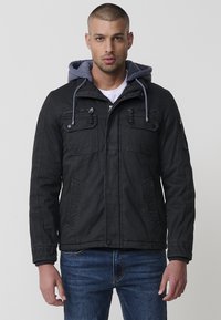 Melnā kapuces jaka ar pelēku oderi, priekšējais rāvējslēdzis, vairāki kabatas un ribotās aproces. Izgatavota no izturīga auduma, ar brīvu siluetu.