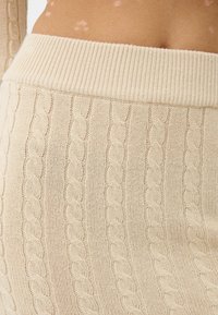 Béžová sukně pletená do kabelového vzoru s širokým žebrovaným pasem. Tkanina má texturovaný vzor propletených kabelů a jemný povrch.