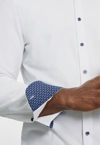 Weißes Hemd mit kontrastierenden blauen Mustern an den Manschetten, mit Knopfverschluss und glatter Textur. Die Manschette zeigt ein geometrisches Design.