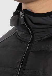 Schwarze Pufferjacke mit hohem Kragen, Reißverschluss und reflektierenden Akzenten. Das Material hat eine glatte Textur mit durchbrochenen Details.