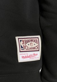 Le tissu noir présente une étiquette tissée "Hardwood Classics" avec des graphismes multicolores, incluant une silhouette de joueur et un motif de basket-ball.