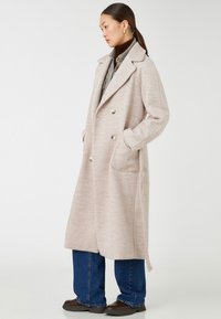 Helle beige lange Wollmischung Jacke mit einem doppelt knöpfbaren Design, großen Revers, seitlichen Taschen und einer sanften Textur. Getragen über einem dunklen Oberteil und Jeans.