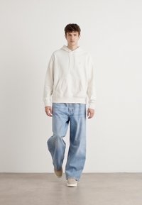 Hoodie bianco in cotone con tasca frontale, abbinato a jeans in denim a gamba larga di un blu chiaro. Modello in piedi su uno sfondo neutro.