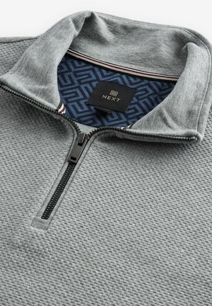 Grauer Reißverschluss-Sweatshirt aus strukturiertem Stoff, grauem Kragen und einem marineblauen gemusterten Futter. Verfügt über einen schwarzen Reißverschluss und ein Logopatch.