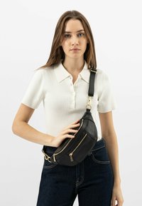 Marsupio in pelle nera con due tasche con zip, cinturino regolabile e accessori in tonalità oro. Indossato a tracolla, superficie texturizzata.
