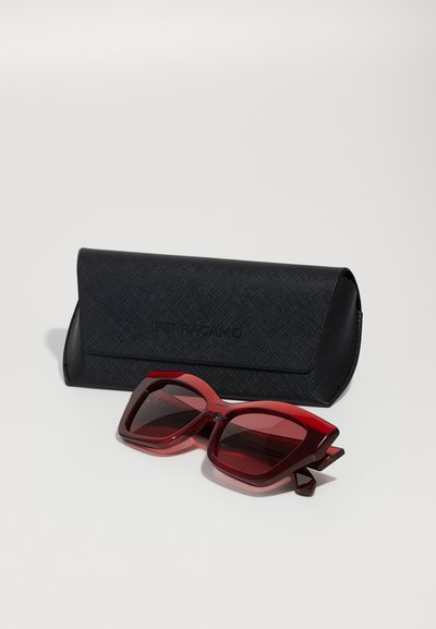 Gafas de sol rojas de gran tamaño con montura transparente y lentes oscuros junto a un estuche negro texturizado con un logotipo en relieve.