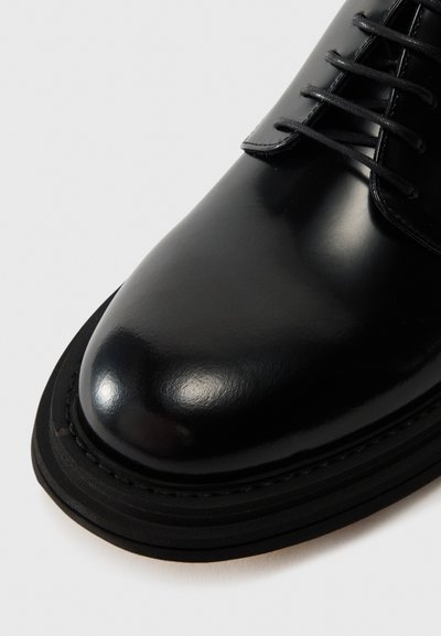 Black leather dress shoe featuring a polished surface, rounded toe, and black laces. The sole has a layered design with stitching details.