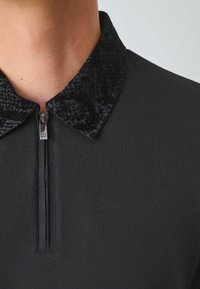 Polo de cuello con cremallera negra que presenta un cuello texturizado negro y un cuerpo liso. El cuello incluye un patrón sutil.