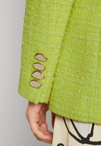 Groene geweven blazer met een gestructureerde stof. Heeft vier accenten van goudkleurige knopen op de mouwen en een subtiel patroon.