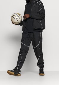 Chándal deportivo negro con acentos blancos, compuesto por una chaqueta ligera y pantalones ajustados. Sosteniendo un balón de baloncesto beige con un diseño en dorado.