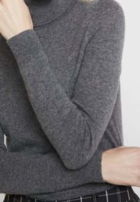 Gray turtleneck sweater in a soft knit fabric. Features long sleeves and ribbed cuffs. Worn with patterned black and white bottoms.
