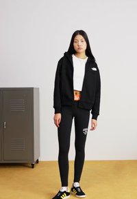 Čierna mikina na zips, biele skrátené tričko, priliehavé čierne legíny, čierno žlté tenisky. Výrazné logo na oblečení. Jednoduché pozadie.