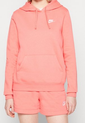 Persoon die een koraalroze Nike hoodie en bijpassende korte broek met witte Nike-logo's draagt, staand tegen een effen achtergrond.