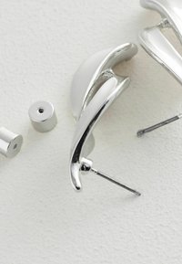 Sudraba krāsas auskari ar elegantu, izliekta dizaina formu. Iekļauj gludu virsmu un pulētas akcentus. Ietver divus metāla aizmugurējos ieliktņus.