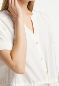 Weiße, kurzärmelige Bluse mit V-Ausschnitt, ausgestattet mit einem Kordelzug an der Taille und perlenartigen Knöpfen auf der Vorderseite. Glatte Stofftextur.