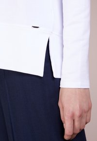 Pohled zblízka na ruku osoby vedle bílé halenky s dlouhým rukávem a bočním rozparkem, spárované s tmavě modrými texturovanými kalhotami.