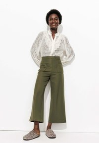 Blouse en dentelle blanche avec des détails froncés, pantalons wide-leg taille haute en olive, et chaussures à imprimé animal à enfiler. Textures lisses, coupe décontractée.