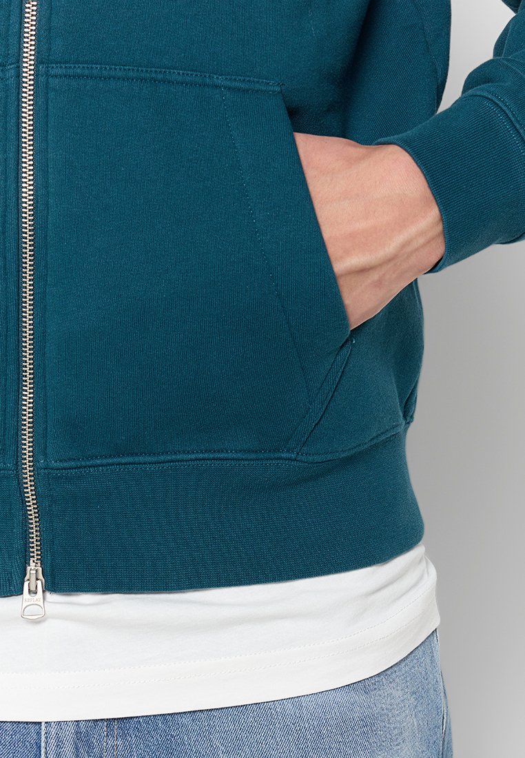 Tee-farbener Zip-Hoodie aus strukturiertem Stoff mit Seitentaschen und einem weißen T-Shirt darunter. Ausgestattet mit einem silbernen Reißverschluss und gerippten Bündchen.