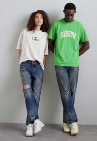 Deux personnes se tiennent côte à côte. L'un porte un t-shirt beige avec un graphique noir et un jean bleu déchiré. L'autre porte un t-shirt vert et un jean bleu délavé. Tous deux portent des baskets blanches.