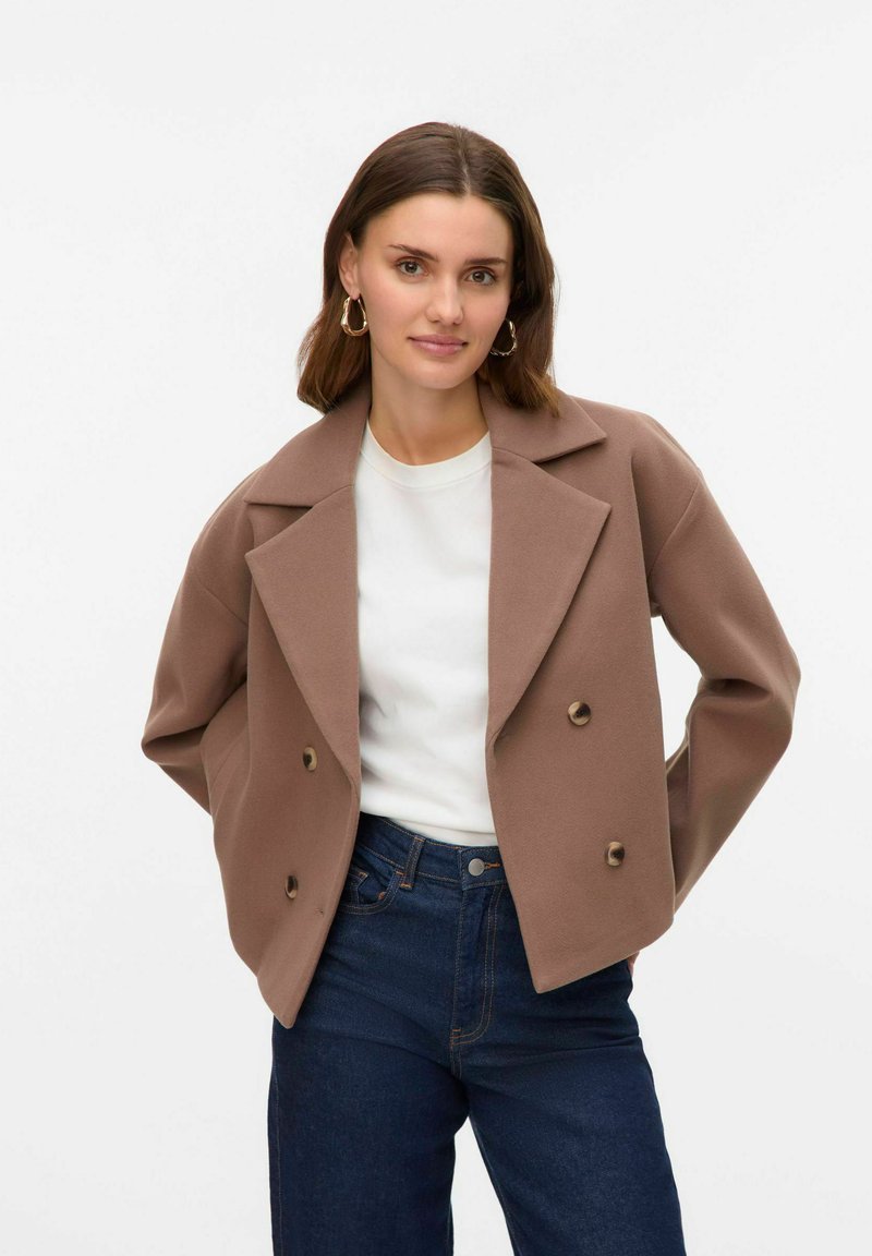 Brun dubbelknäppt jacka med breda slag, bärs över en vit skjorta och mörka jeans. Materialet verkar mjukt och texturerat, med runda knappar.