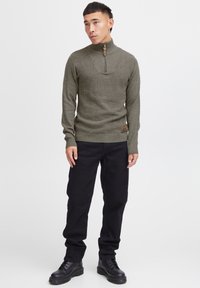 Olivgrüner gerippter Strickpullover mit hohem Kragen und Reißverschluss, versehen mit einem braunen Patch am Saum. Kombiniert mit schwarzen Hosen und Stiefeln.