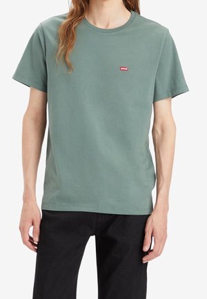 Personne portant un t-shirt Levi's à manches courtes vert kaki avec un petit logo rouge sur le côté gauche de la poitrine et un pantalon noir.