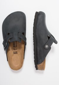 Sabots en caoutchouc noir avec boucle ajustable, semelle en liège et semelle extérieure texturée noire. Marquage latéral visible, design à dos ouvert. Taille 37.