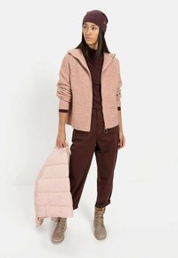 Lichtroze ribgebreide cardigan, bordeaux turtleneck, bruine broek, beige laarzen, houdt een lichtroze pufferjack vast. Eenvoudig, functioneel ontwerp.