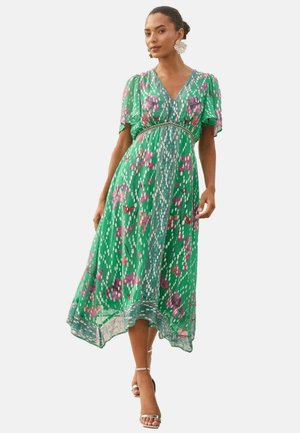 Frau trägt ein grünes Midikleid mit Blumenmuster, kurzen, flatternden Ärmeln und V-Ausschnitt, kombiniert mit silbernen Riemchenabsätzen und großen Ohrringen.