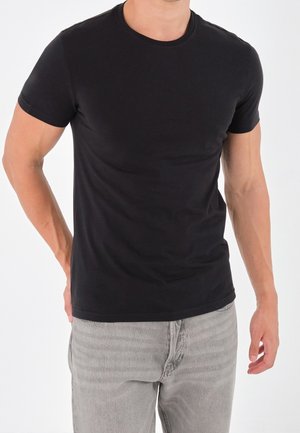 Man draagt een nauwsluitend, effen zwart T-shirt met korte mouwen en lichtgrijze spijkerbroek, staand tegen een witte achtergrond.