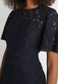 Marinblå spetsklänning med korta fladdrande ärmar, ett blommönster och en texturerad midja. Tyget har en fin, invecklad design.