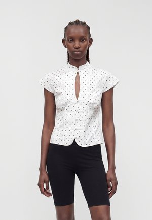 Femme portant un chemisier blanc à manches courtes avec des pois noirs et une ouverture frontale, associé à un short noir ajusté, debout devant un fond uni.