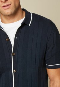 T-paita lyhyillä hihoilla, tummansininen valkoisilla yksityiskohdilla, jossa on ribbikuvio, pystyraitoja, nappikiinnitys ja klassinen kaulus.