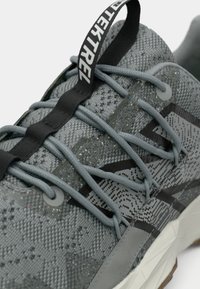 Grauer Sportschuh mit oberer Schicht aus Textilstruktur, schwarzen Akzentlinien und gestreiftem Absatz. Verfügt über runde Schnürsenkel und ein Logo auf der Fersenschlaufe.