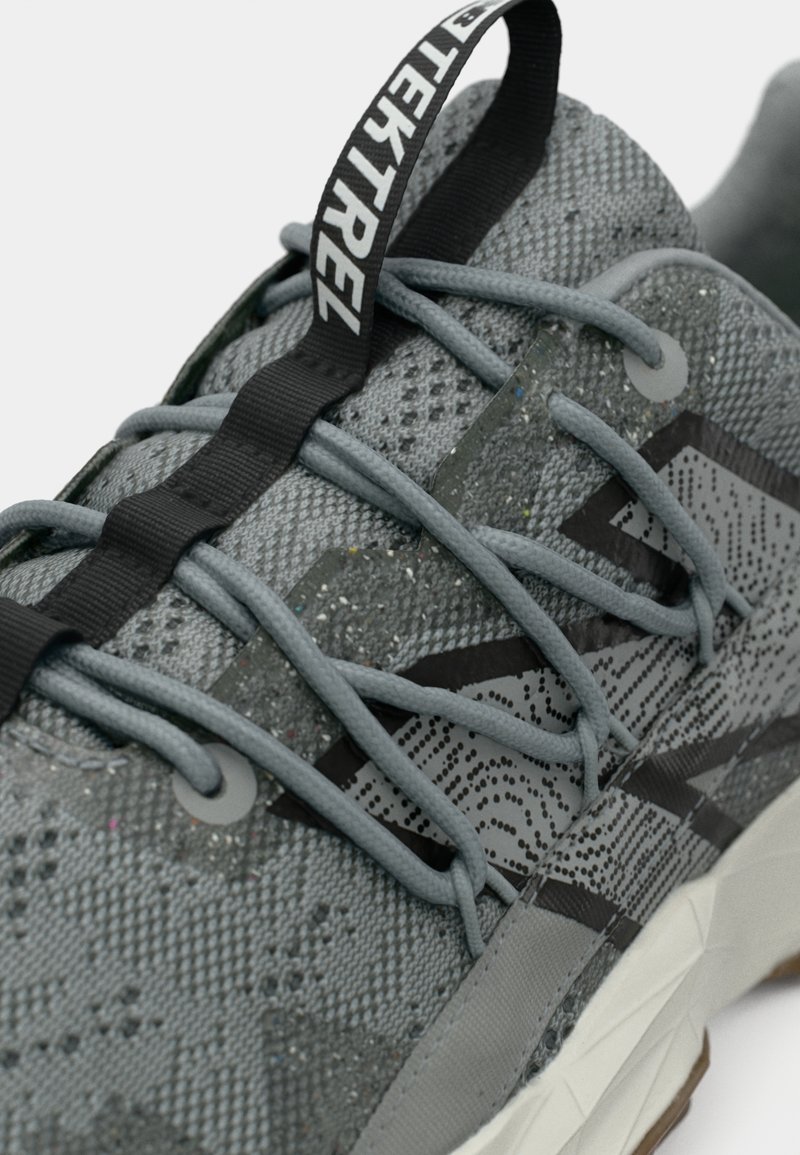 Scarpa sportiva grigia con tomaia testurizzata, linee di accento nere e un tallone a strisce. Presenta lacci arrotondati e un logo sulla linguetta del tallone.