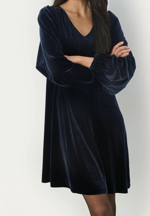 Vrouw die een losse marineblauwe velvet jurk met lange ballonmouwen draagt, staat met gekruiste armen tegen een effen witte achtergrond.