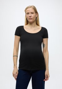 Trudna žena s dugom plavom kosom koja nosi crnu kratku majicu i tamnoplave hla če, stojeći protiv jednostavne svijetle pozadine.