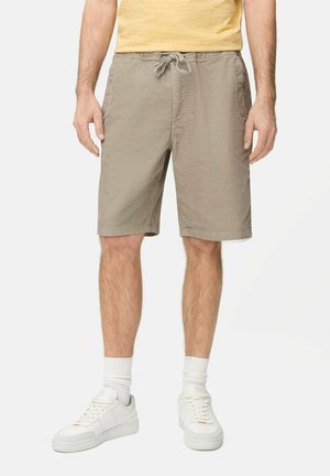 Mężczyzna ubrany w beżowe szorty z troczkami, białe skarpetki i białe trampki, stojący na tle jednolitego tła.