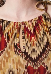 Multifarbener gemusterter Oberteil mit Rundhalsausschnitt und Schlüssellochdetail. Der Stoff ist leicht und hat eine glatte Textur; die Farben umfassen Rot, Braun und Gelb.
