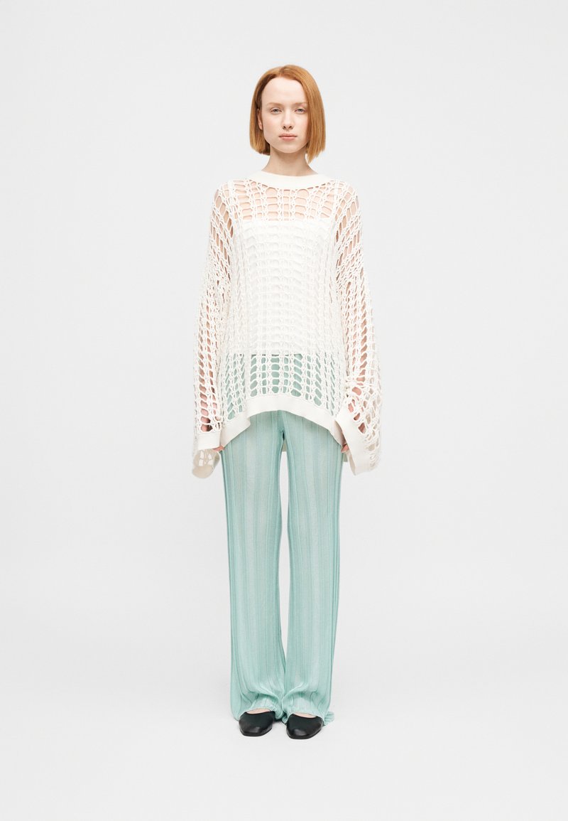Λευκό πλεκτό πουλόβερ με ανοιχτό πλέγμα, συνδυασμένο με πλισέ παντελόνια σε μέντα πράσινο χρώμα. Μαύρα παπούτσια συμπληρώνουν την εμφάνιση.