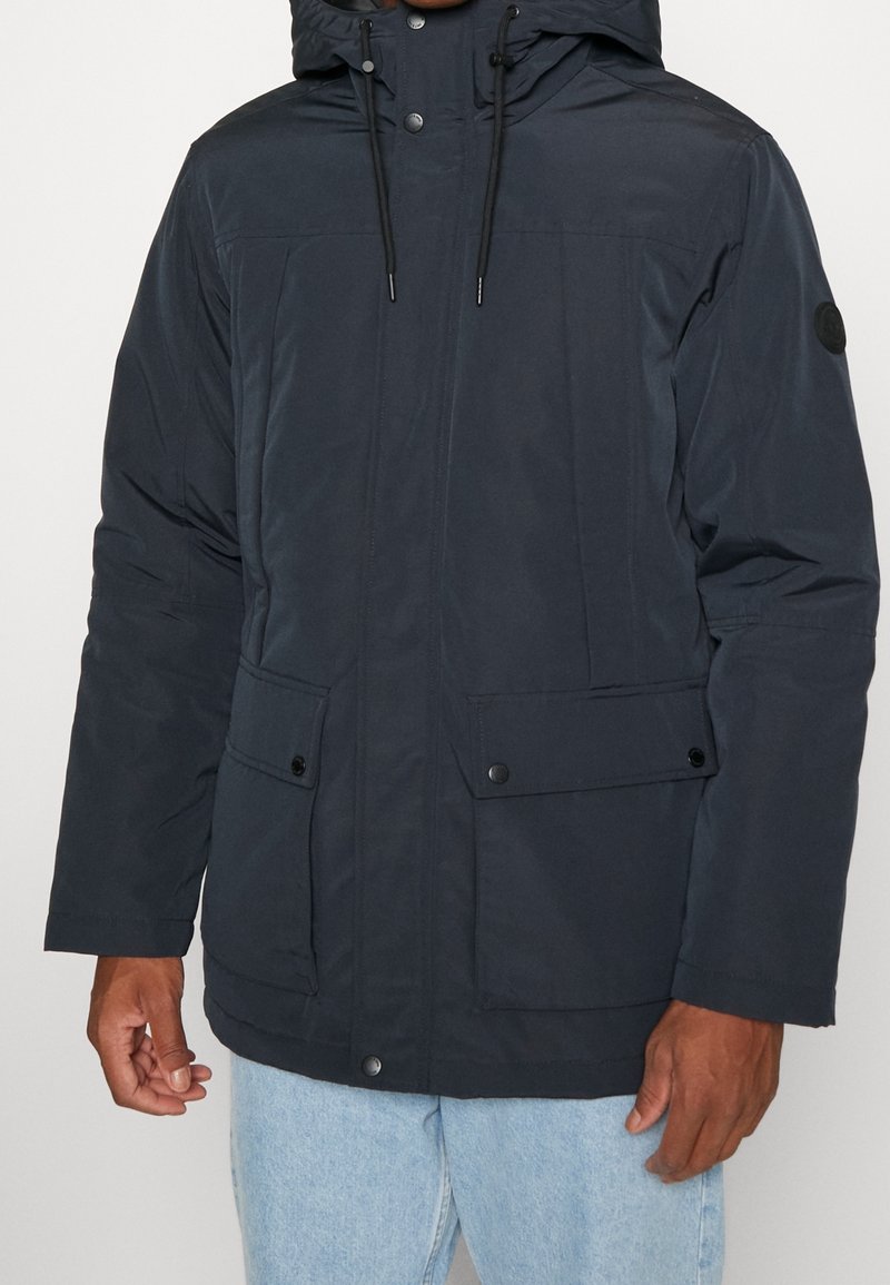 Man draagt een donkerblauwe parka met voorvakken met kleppen, drukknopen en verstelbare koordjes aan de capuchon, gecombineerd met lichtblauwe spijkerbroek.