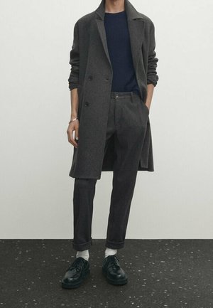Person wearing a dark gray long coat, navy sweater, dark gray pants with cuffs, white socks, and black dress shoes standing against a plain background.