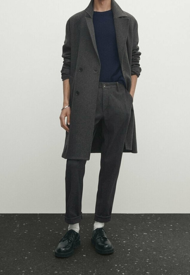 Personne portant un long manteau gris foncé, un pull bleu marine, un pantalon gris foncé avec des revers, des chaussettes blanches et des chaussures habillées noires, debout devant un fond uni.
