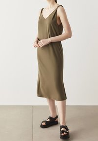 Robe sans manches vert olive avec un décolleté en V profond et des détails de bretelles, associée à des sandales noires avec des accents dorés et des sangles texturées.